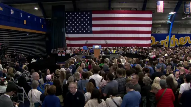 A venue with a packed crowd looking ahead at an empty stage with a podium that says "Harris-Walz"