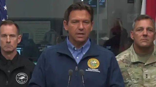 Florida governor Ron DeSantis soeaking behind two microphones. He is wearing a dark blue jacket and shirt, slightly lighter in colour. Two men stand behind, one in black and one in military fatigues.