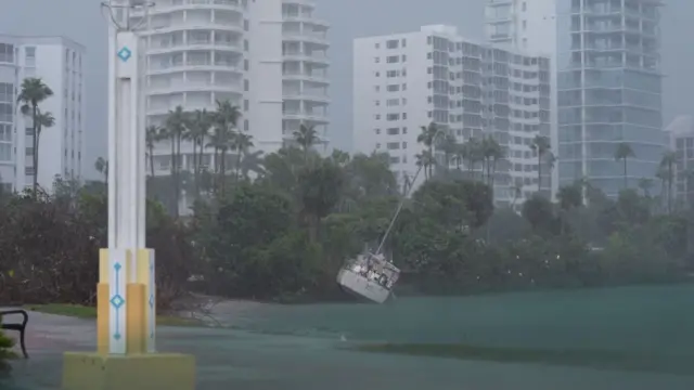 A boat is washed up into some trees as a result of Hurricane Milton. The trrees are in front of several white residential buildings. A white and yellow pole is in the foreground