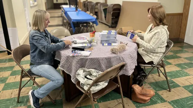 Two white blonde women sit on folding chairs at a table full of brochures
