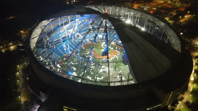 Ariel photo of a baseball stadium with a destroyed roof. The field is clearly visible as the floodlights are on. There are many parts of the roof scattered across the playing service.