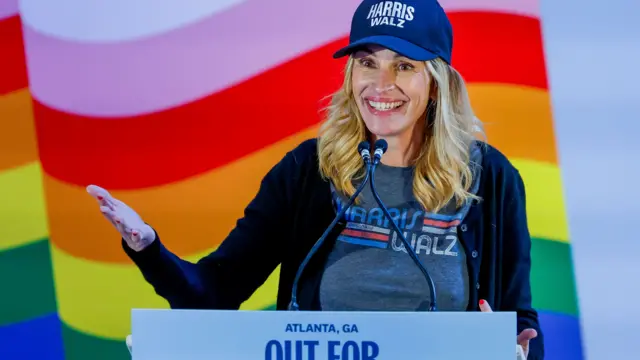 Actress Julia Roberts, wearing a Harris and Walz T-shirt and cap, stands in front of a pride flag and addresses a crowd in Georgia