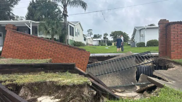 Brown brick wall, a metal fence, and pieces of tarmac crumbled down into the ground. A number of homes and palm trees can be seen in the background, behind the wall