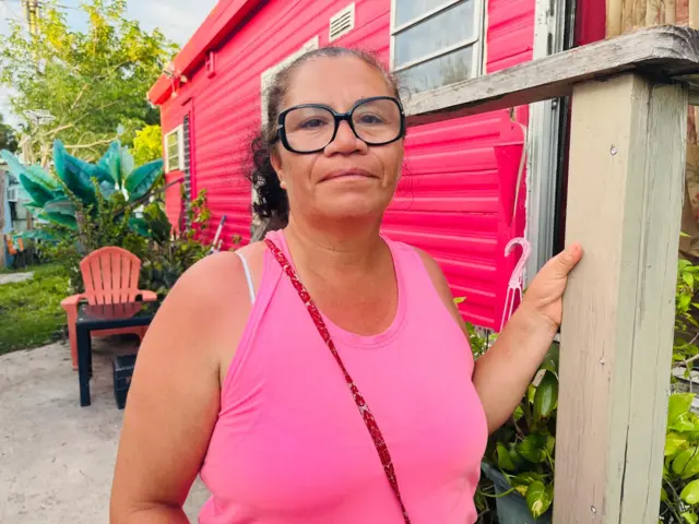 Maria looks into the camera in a pink shirt outside her pink home