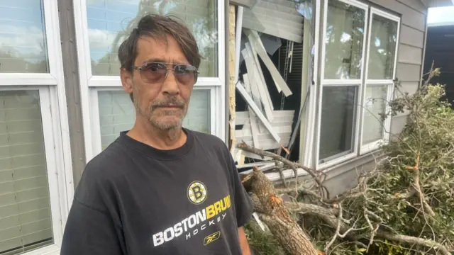 Man wearing dark sunglasses looks at camera. Behind him is house which has had window broken by a big tree branch, that is now resting on the floor