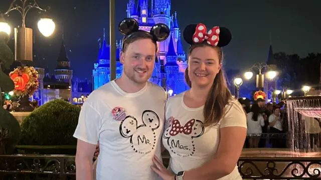 Jennie Flin and her spouse, wearing Mickey and Minnie Mouse Ears, pose in Disney World, Florida