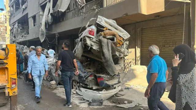 Cars and rubble piled up on the side of a small street with people walking