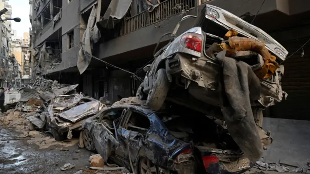 Damaged cars line the side of a small street. Some are piled on top of each other