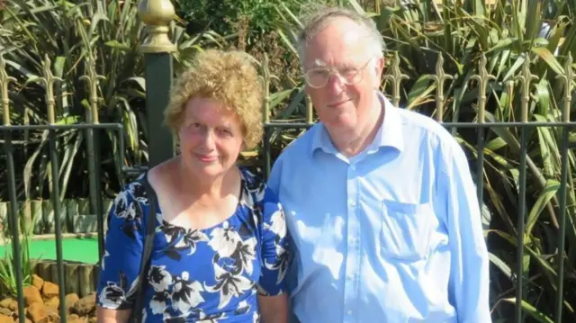 Lois and John McCullough, standing in front of a mini golf course. They are both wearing blue and smiling at the camera.