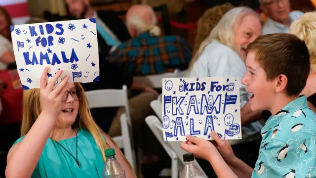 Children, a boy and girl, hold up signs which read "Kids for Kamala"