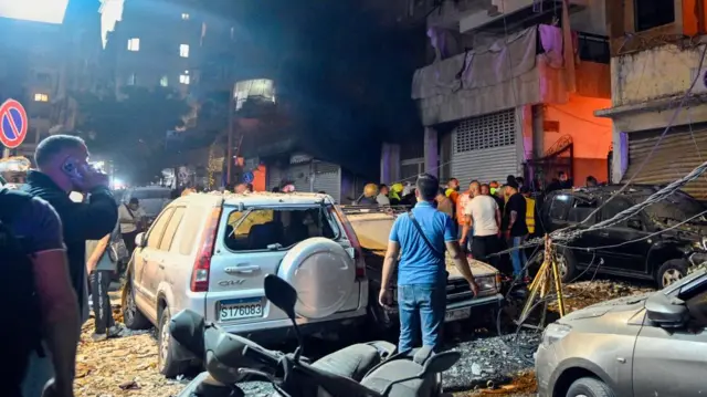 A group of people look at damaged cars following an Israeli airstrike on Beirut. To the left of the image, a man speaks on a mobile phone.