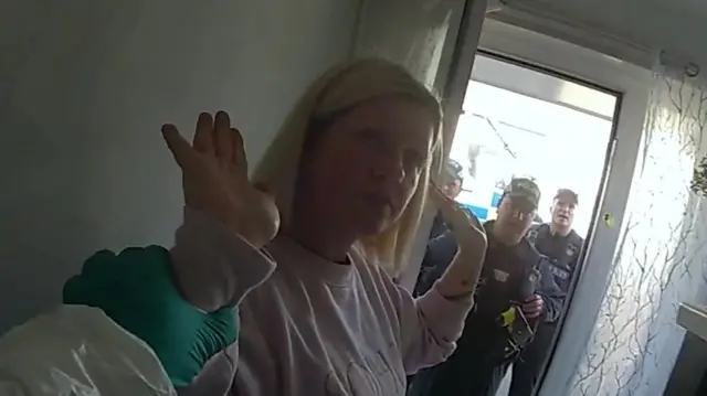 Virginia McCullough raises her hands in a surrender motion as police surround her. She is wearing a pink jumper