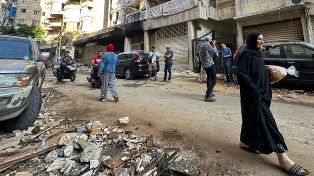 Image of a damaged street in Beirut. A woman on the right carries bread as several people behind her look on. At the rear of thew shot, two men are on mopeds.