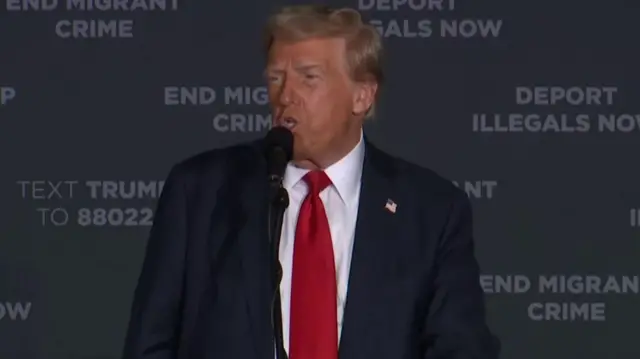 Trump, wearing a blue suit and red tie, speaking into a microphone in front of a board that says "end migrant crime, depart illegals now"