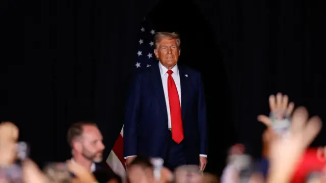 Donald Trump, wearing a blue suit and red tie, grimaces while on stage in Aurora, Colorado