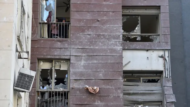 The outside of a building where the glass windows have been smashed. Some people stand in a room in the top right