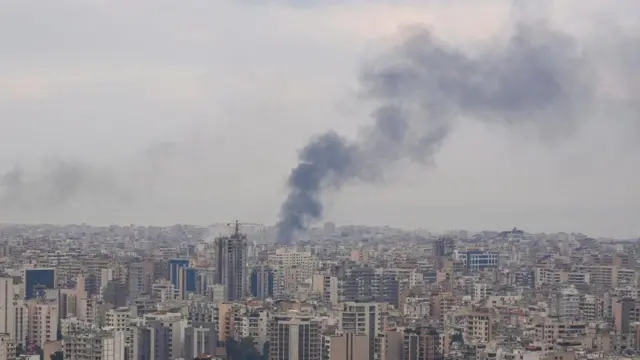 A plume of smoke raising high in the air over Beirut. Wide shot of the city's skyline, with the smoke billowing from the centre of the picture.