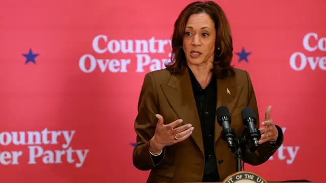 Kamala Harris, wearing a khaki suit and black shirt, gestures with her hands as she speaks into a microphone in front of a pink background