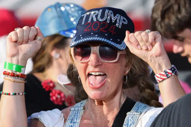 Woman at Coachella rally raises both fists. She is wearing a sequined Trump 2024 hat