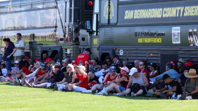 Dozens of people sitting and waiting in the shade next to of San Bernadino County Sherriff buses