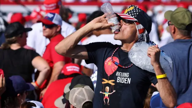 A man holds a bag of ice on his shoulder and is drinking a bottle of water. He is wearing a tshirt that reads "Beijing Biden" and has an image of Joe biden as a marionette, which has a Chinese flag baloon attached to it.