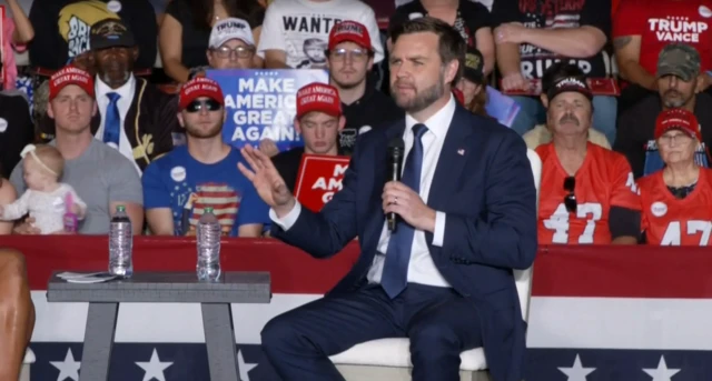 JD Vance speaking at a town hall-style rally, he's sitting at a table while supporters watch in the background