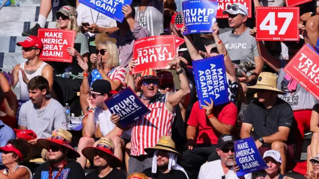 Trump supporters sitting in the stand holding signs which read "Make America Great again" , "47" and "Trump Vance"