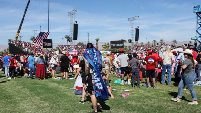 A crowd of hundreds of people waiting at the rally. There are signs which read "Better off with Trup" and many of them are wearing hats to keep off the sun.