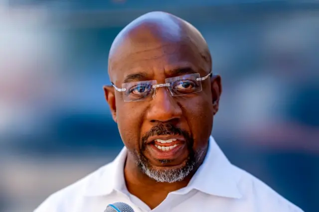 Democratic Senator Raphael Warnock. He is wearing a white shirt and rimless glasses and looking just off camera.