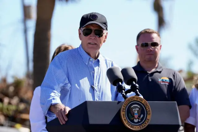 President Biden giving a press conference while visiting areas affected by Hurricane Milton in Florida. He's wearing a dark cap and aviators and standing behind a podium.