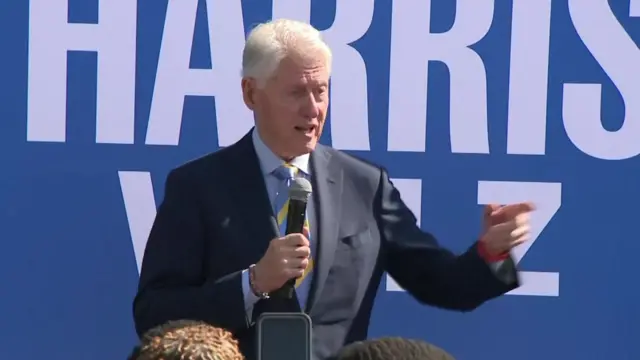 Bill Clinton standing in front of a blue Harris Walz campaign sign