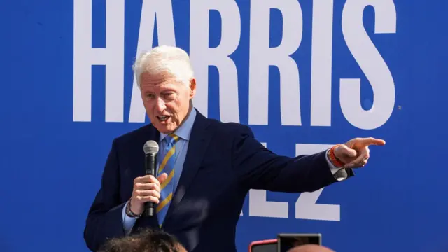 Bill Clinton, standing in front of a Harris-Walz sign, points to the left as he addresses a crowd