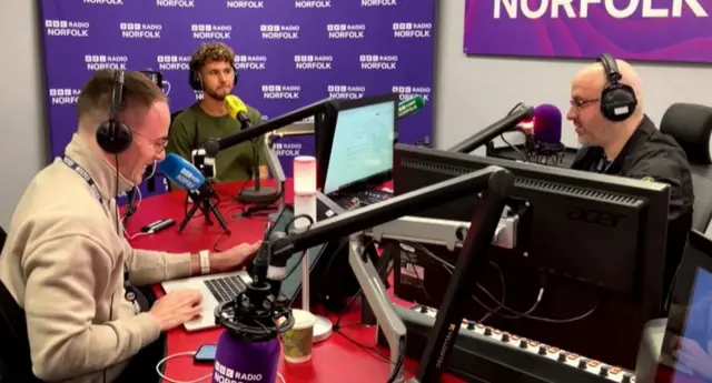 Emiliano Marcondes sits in a radio studio with two people wearing headphones