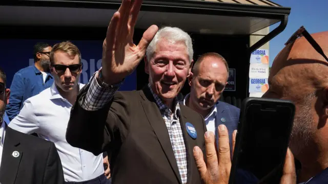 Bill Clinton waves towards a camera while wearing a blue Harris-Walz badge on his jacket