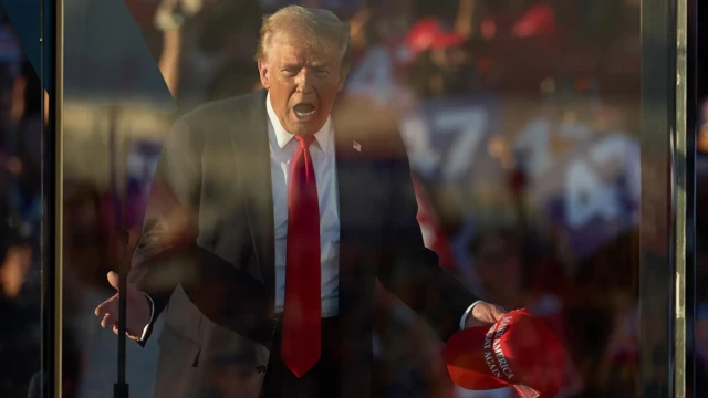 Trump gestures with his hands out as he speaks at the rally holding a red MAGA cap. Light catches on the protective glass between Trump and the audience