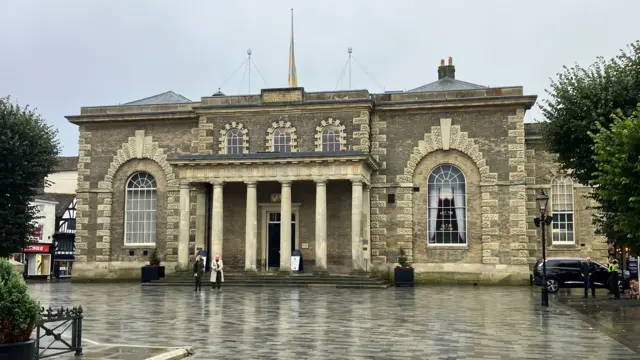 Salisbury Cathedral on a cloudy, rainy day. Two people are standing outside of the guildhall and a police officer can be seen standing next to a car on the right