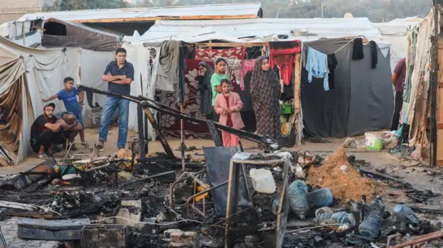 Seven people look on at damage caused in Gaza by an Israeli airstrike. In the front of the picture is the damage with the people in the middle.There are clothes hung up behind them. To the right of the frame part of a man is visible, he is lookig at damage elsewhere.