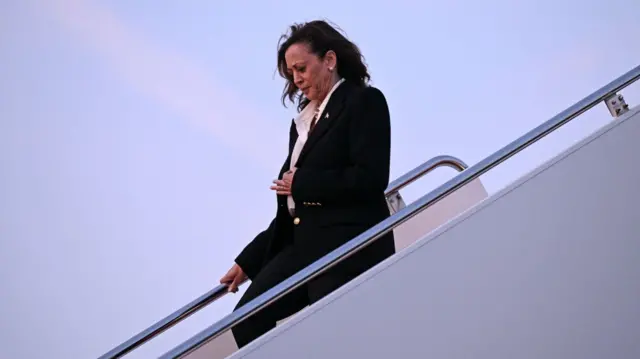 Kamala Harris, wearing a black suit and white shirt, looks down as she disembarks Air Force Two on Sunday