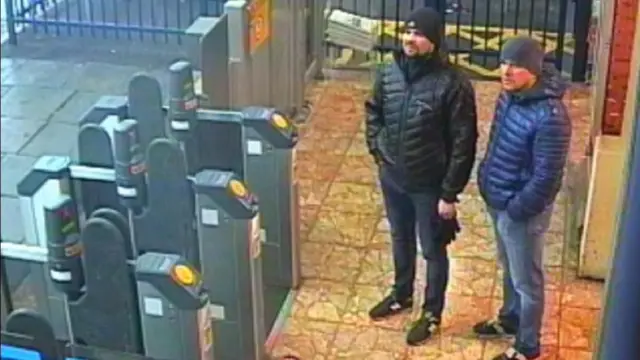 A CCTV image of two men standing by ticket barriers