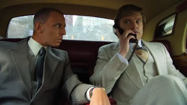 A still from the film shows actor Jeremy Strong, wearing a grey suit, leans towards fellow actor Sebastian Stan, wearing a lighter grey suit, in the back of a car