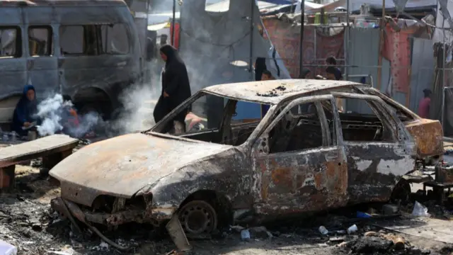 A woman walks behind a burnt-out car