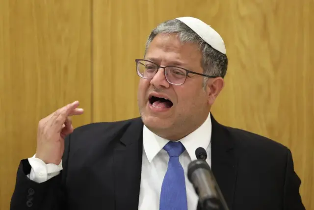 Itamar Ben-Gvir delivers speech in black suit, white shirt and blue patterned tie. His right hand is raised, the tops of his pointer finger and thumb touching, his mouth open