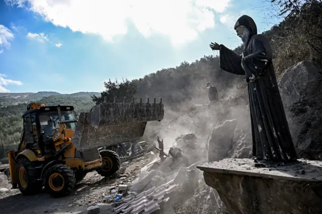 A bulldozer clears rubble by a statue of Charbel, a patron saint of Lebanon