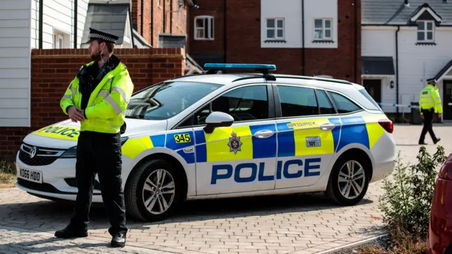 Police officers and police cars outside the Muggleton Road property of Charlie Rowley and Dawn Sturgess. There is a police car visible and police tape