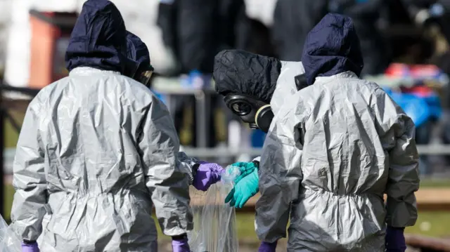 A group of people stand together examining a plastic bag during the novichok poisonings operation in Salisbury. They are wearing black gas masks and grey hazmat overalls