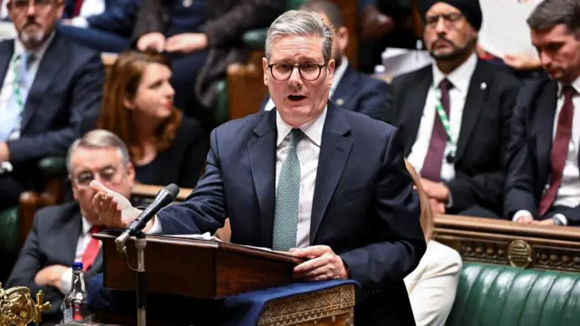 Keir Starmer in a suit with a green tie at the check-in box in the House of Commons, with MPs sitting on benches around him