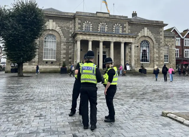 Three police officers standing in front of the Guildhall in Salisbury. A few people walk around in the square behind them