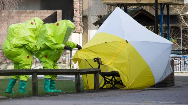 Two people in yellow hazmat suits and head protective gear examine a bench under a forensic tent in Salisbury after the novichok poisoning of the Skripals