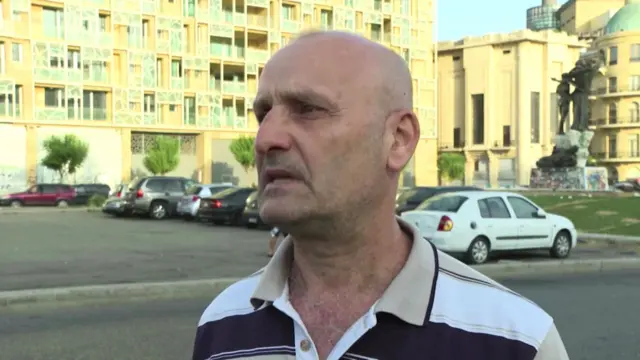 Mohamed Gradi close us. Bald man wearing black, white and beige striped polo shirt. In background is a parking lot with several parked cars, buildings illuminated by early morning sunshine