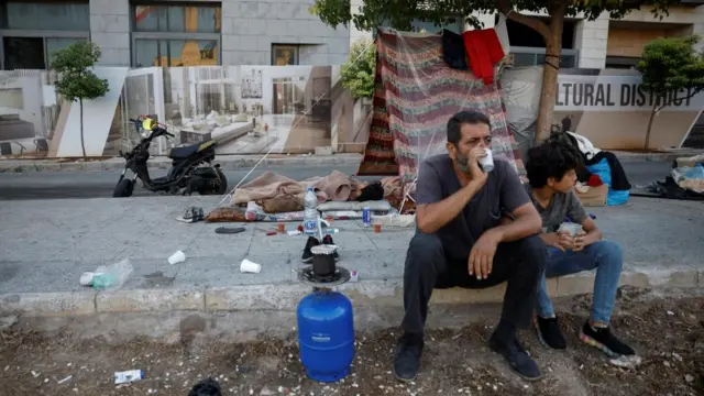 A man and a boy drink from plastic cups while sitting on a curb in front of a makeshift tent on the streets of central Beirut